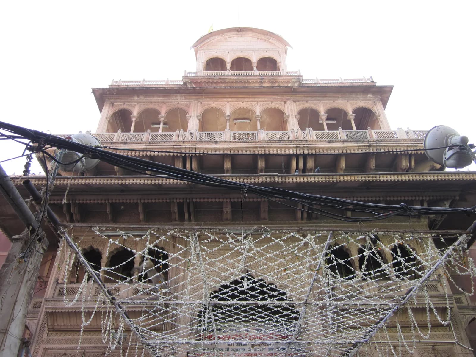 The upper storeys of the Banke Bihari temple in Vrindavan seen from the lane below, arched galleries and a domed pavilion strung with white flower garlands